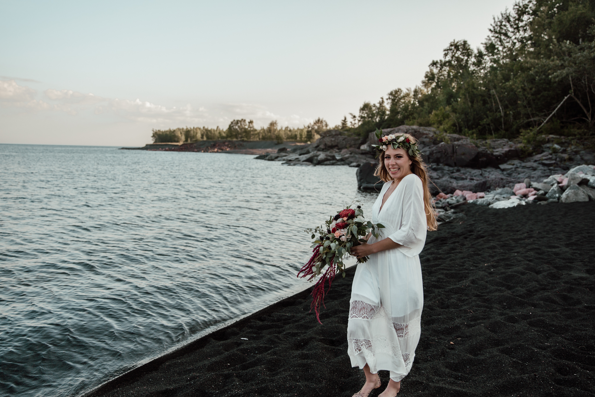 bride in a beach dress holding a bouget giggling in a portrait on a black sand beach next to a lake
