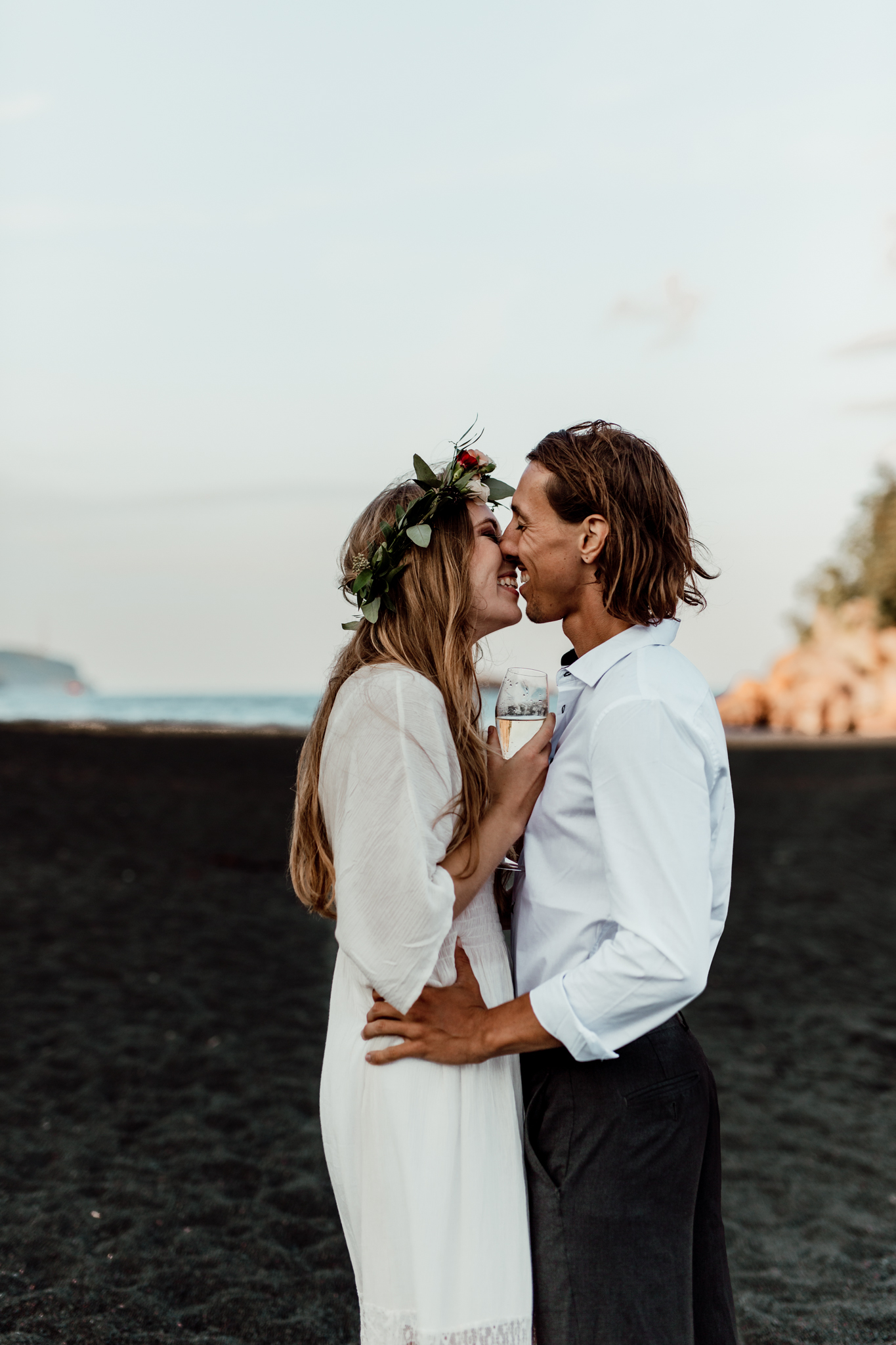 couple going in for a kiss while holding champagne on a black sand beach 