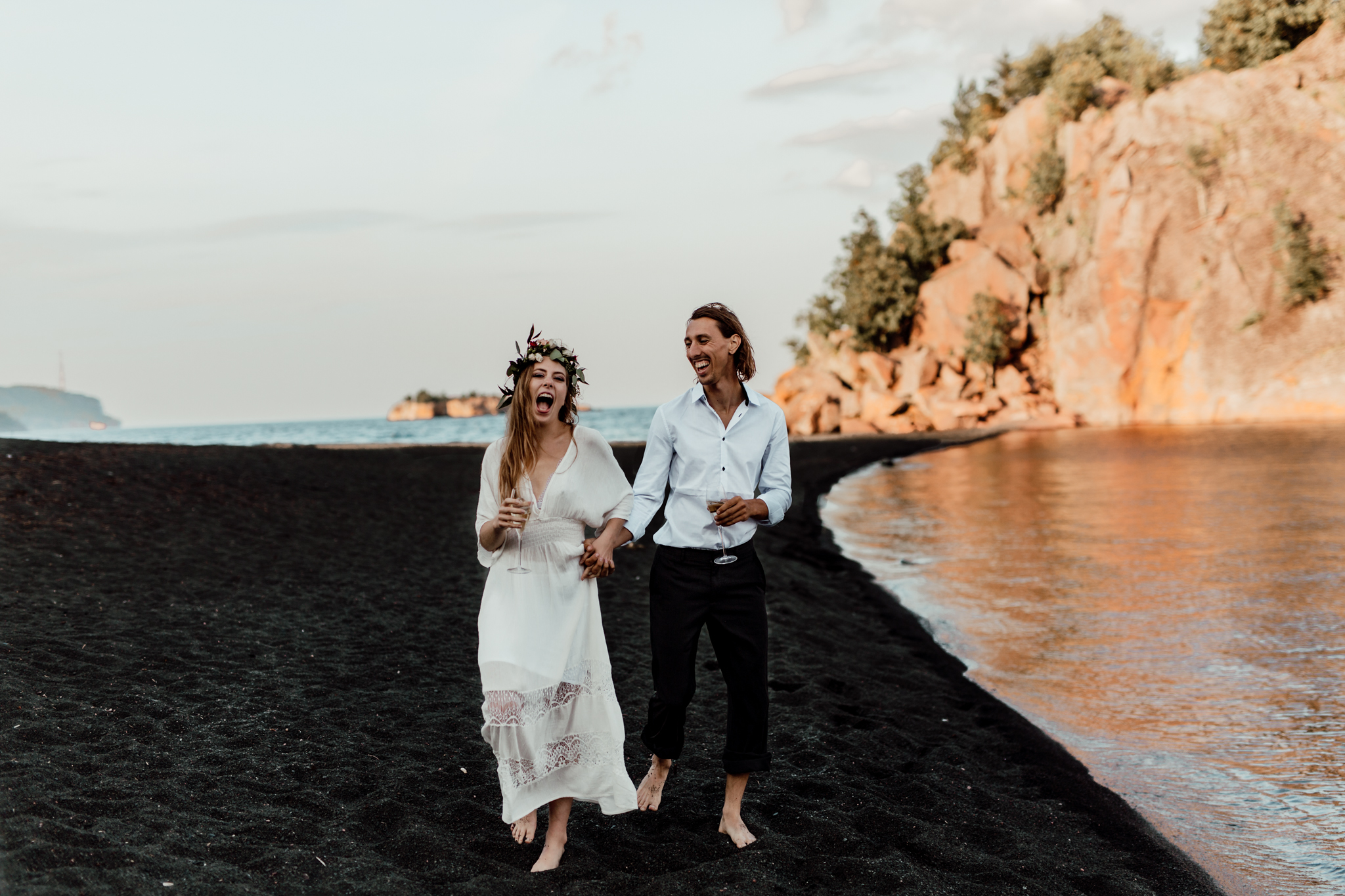 elopement with bride and groom holding champagne while walking on a black sand beach 