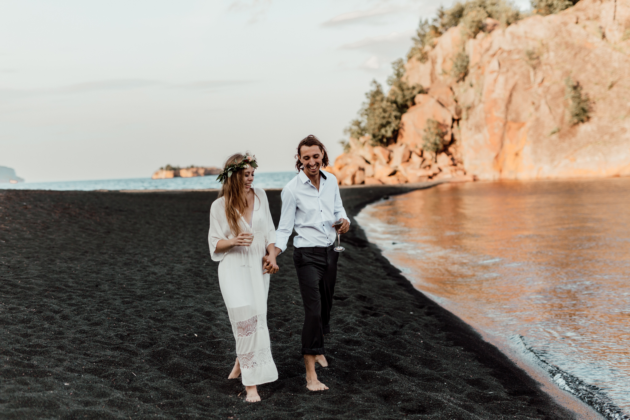elopement with bride and groom holding champagne while walking on a black sand beach 