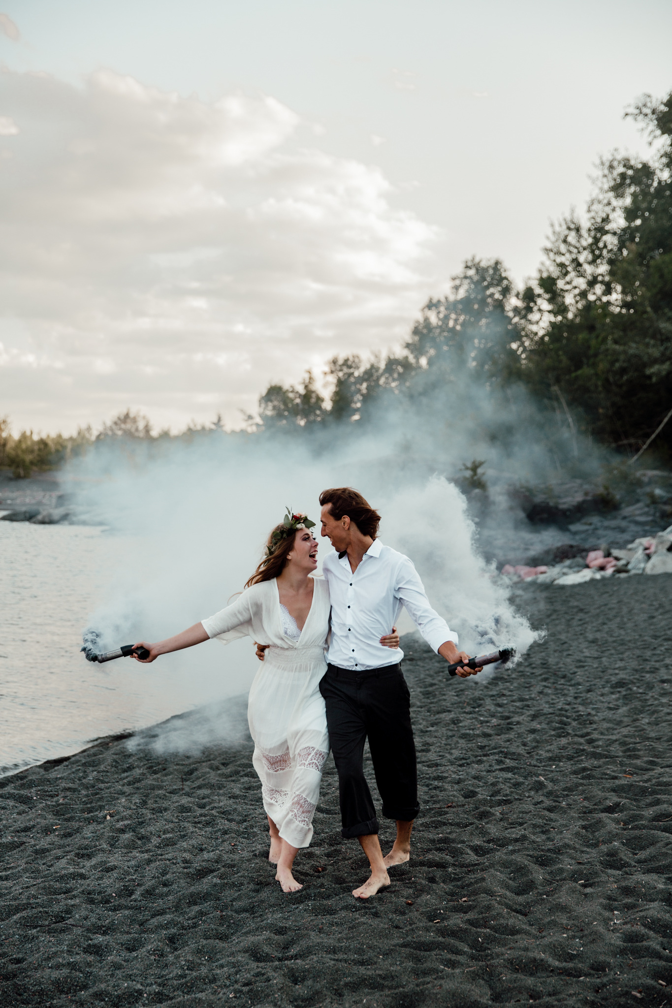 bride and groom walking on a black sand beach holding smoke bombs looking at each other and laughing