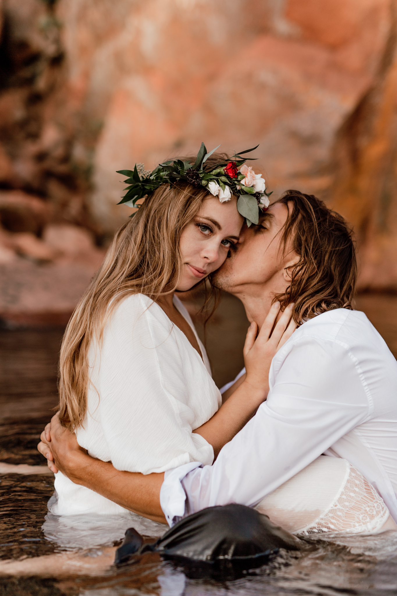elopement portraits with bride looking at the camera as groom kisses her cheek while they sit in a lake 