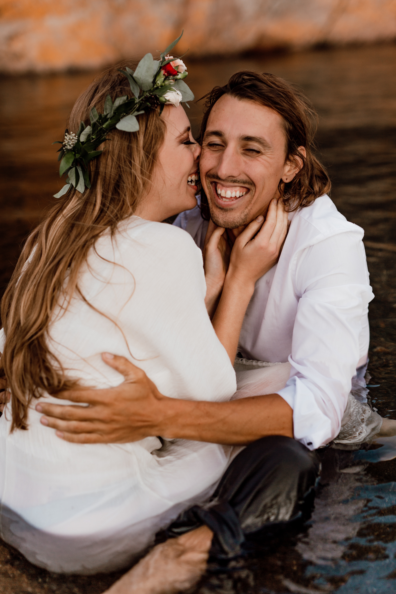 bride and groom sitting in the lake facing each other and holding each others faces 