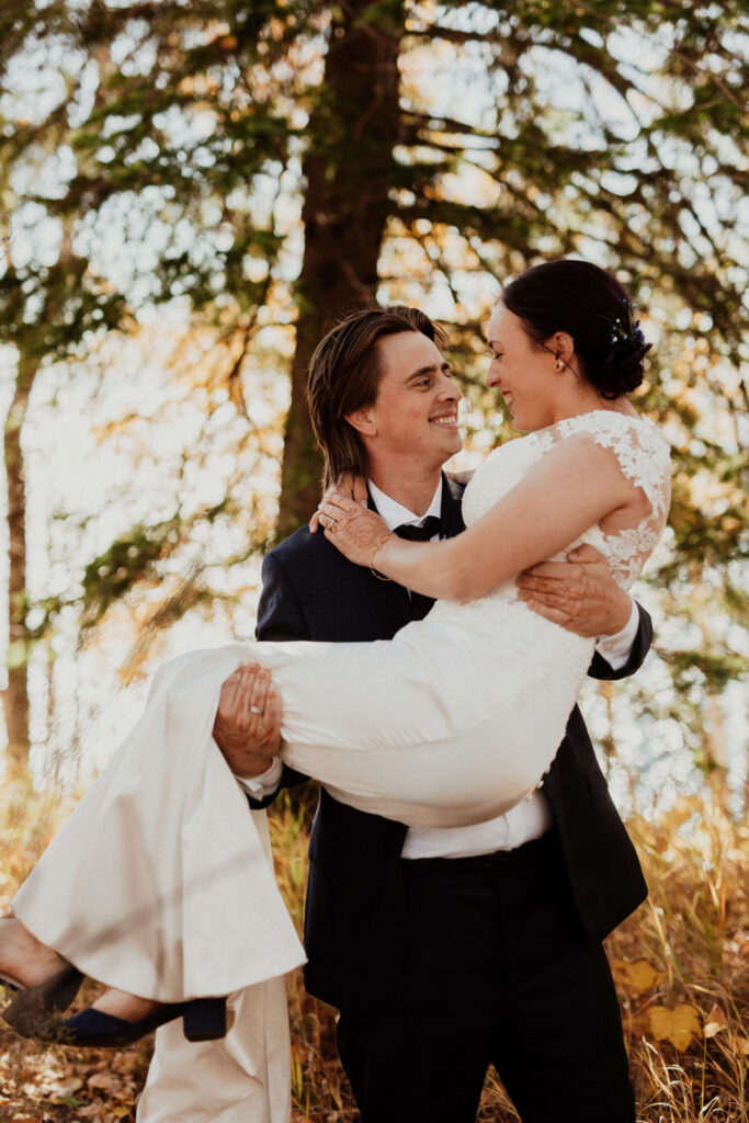 groom picked up bride and carried her with smiles along a backdrop of autumn trees 