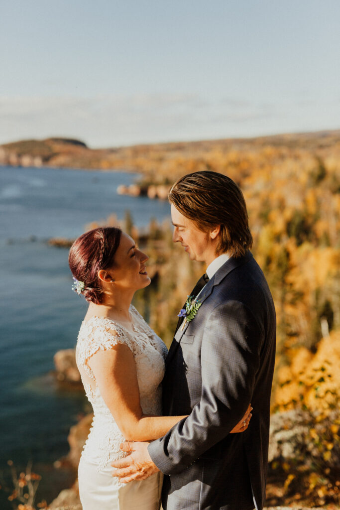 bride and groom holding and looking at each other with minnesota's shoreline behind them and fall colors in full effect 