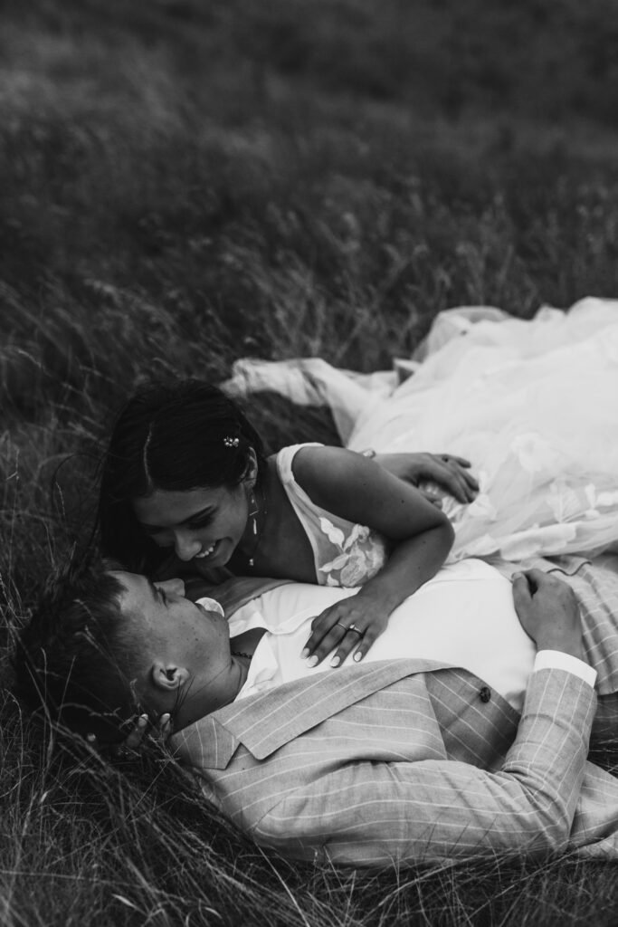 black and white image with couple laying in tall grasses. Bride is giggling laying on grooms chest 