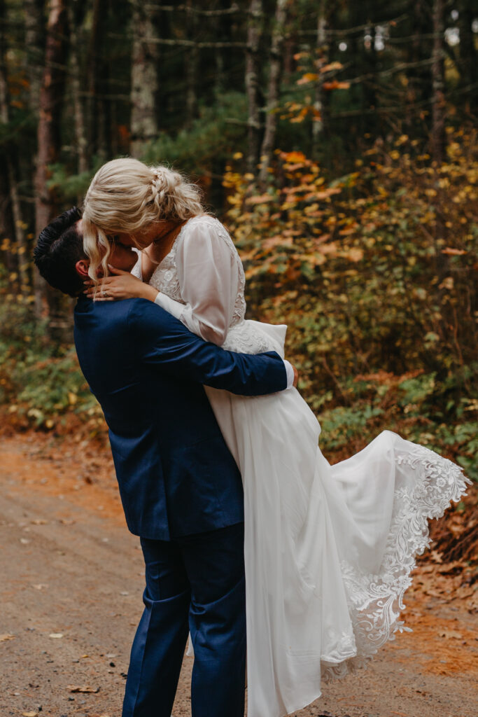 groom picked up bride and are sharing a kiss along a forest road with fall colors in the background