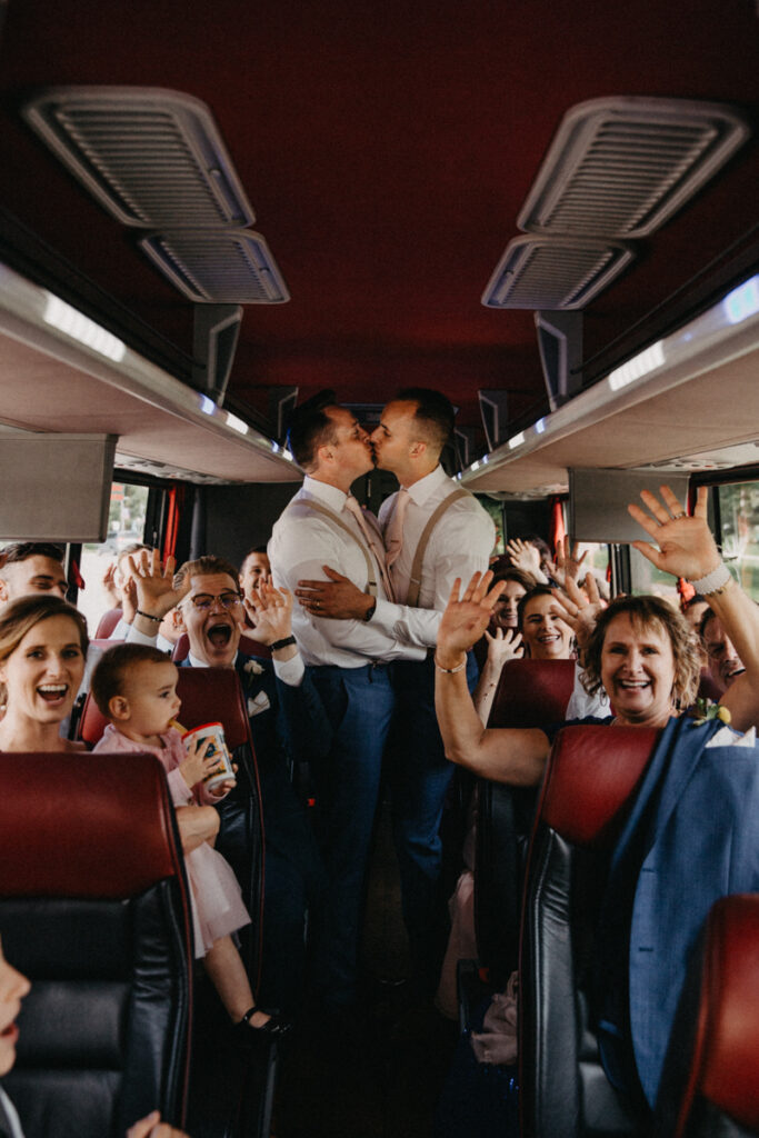 grooms are kissing in the middle of a chartered bus isle where all friends and family are gathered around cheering 