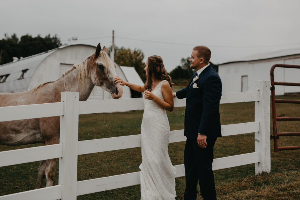bride and groom leaning against a white fence petting a horse on their wedding day 