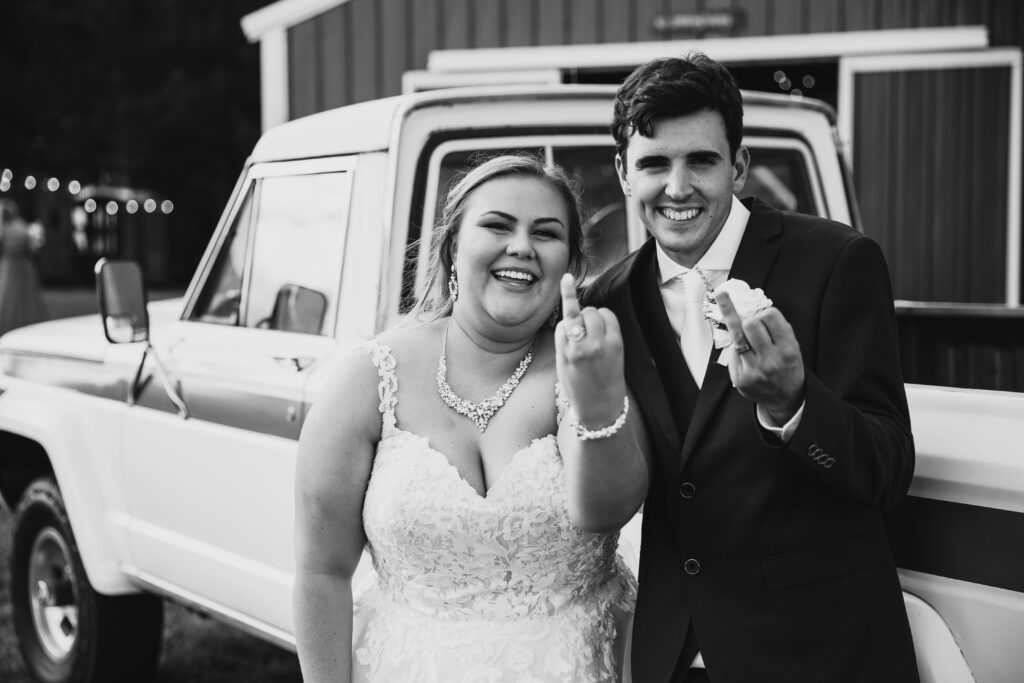 black and white image of bride and groom holding up their ring fingers like their flipping off the camera 