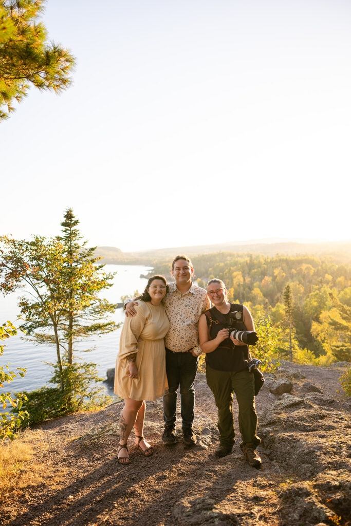 portrait of brianna kirk holding camera smiling with two clients overlooking a cliff at sunset