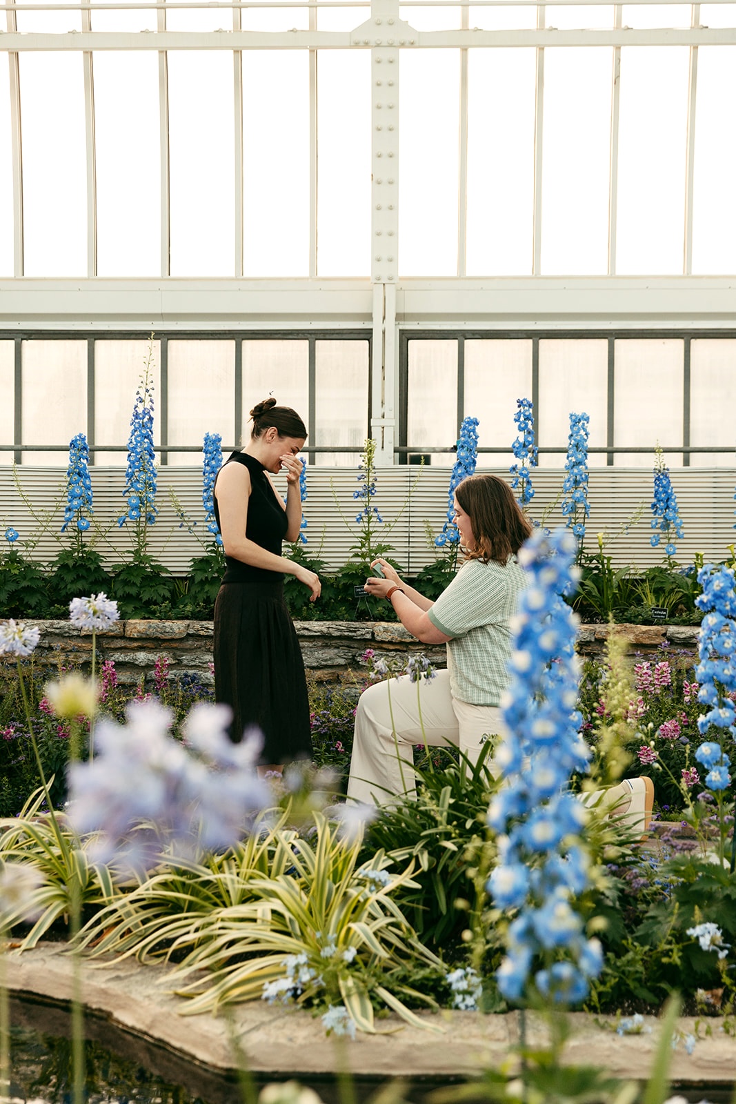 lesbian surprise proposal with the woman on one knee and the other in shock surrounded by blue flowers
