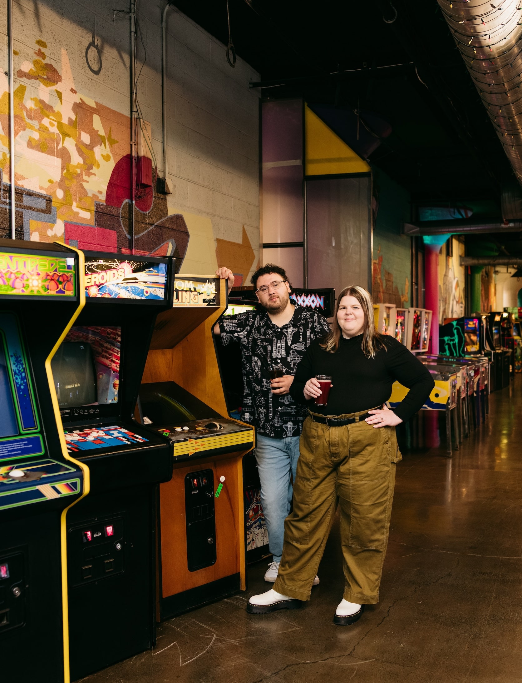 couple stands side by side smiling with vintage arcade games behind them at can can wonderland in minneapolis